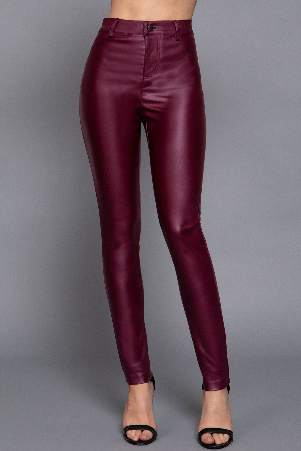 A model's lower body wearing high-waisted, shiny burgundy faux-leather skinny pants and black heeled sandals, photographed against a solid gray studio background.