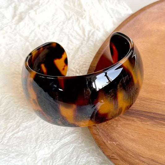 A glossy tortoiseshell cuff bracelet with dark brown, amber, and black mottled patterns, resting on a textured white surface next to a wooden board.