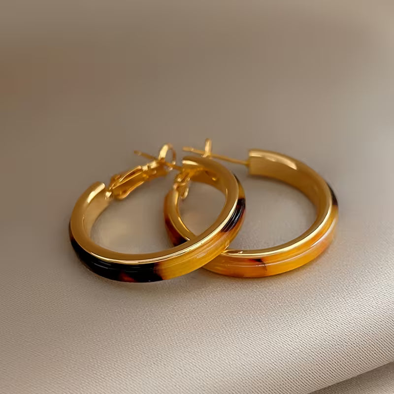 A pair of gold hoop earrings with a glossy amber-yellow and tortoiseshell pattern, resting on a textured beige fabric background.