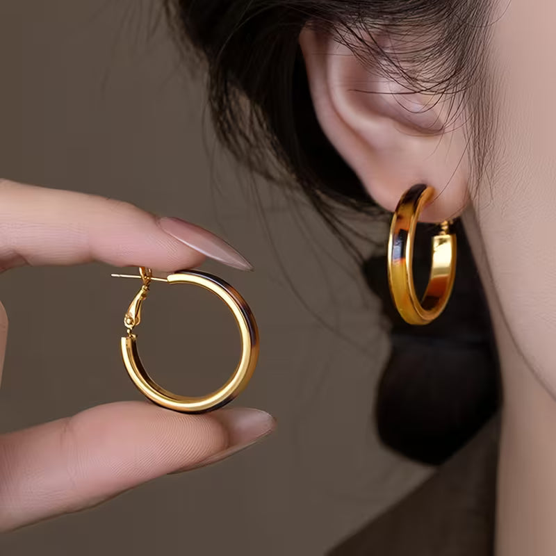 A close-up lifestyle shot of a person wearing a polished gold hoop earring on their ear while holding another matching gold hoop earring in their hand.