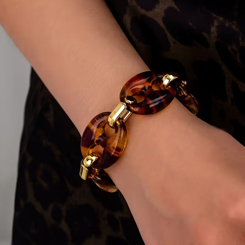 A close-up of a person's wrist wearing a chunky link bracelet made of oval amber-brown translucent links with shiny gold-toned metal connectors, against a dark leopard-print garment.