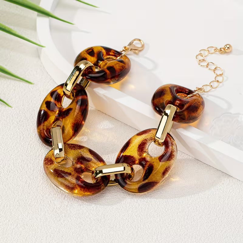 A chunky link bracelet with glossy amber-brown and black leopard print resin links connected by gold-tone metal, displayed on a white surface with a white tray and green leaves as props.