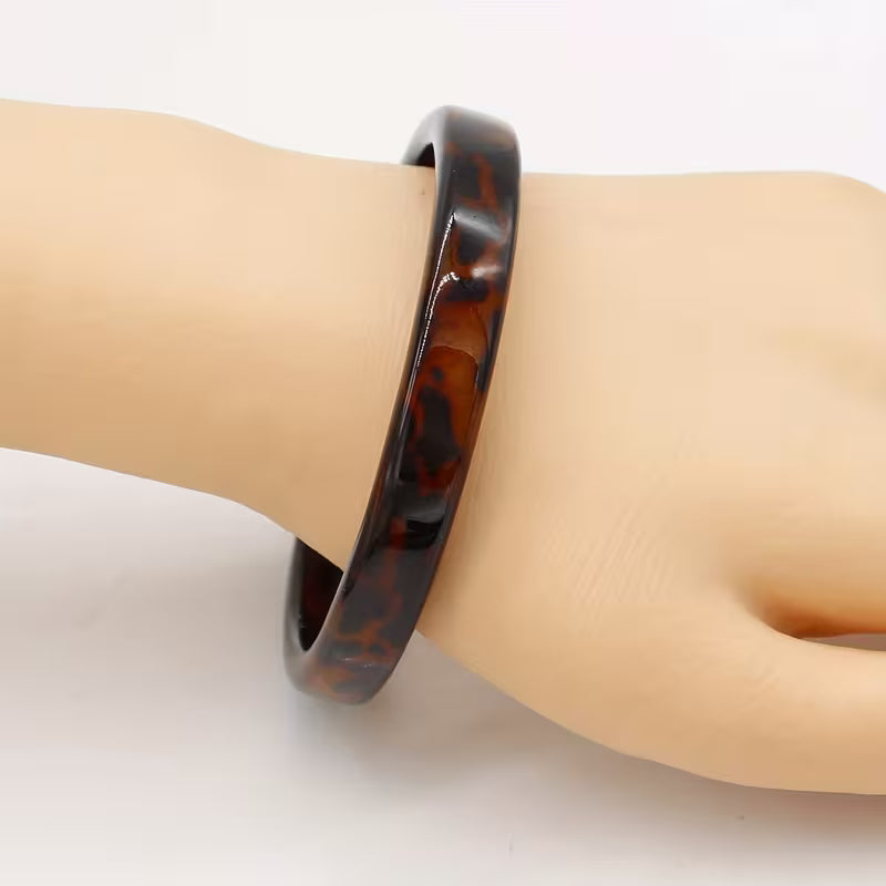 A glossy tortoiseshell bangle bracelet with dark brown and amber swirls, shown worn on a beige mannequin hand against a plain white background.