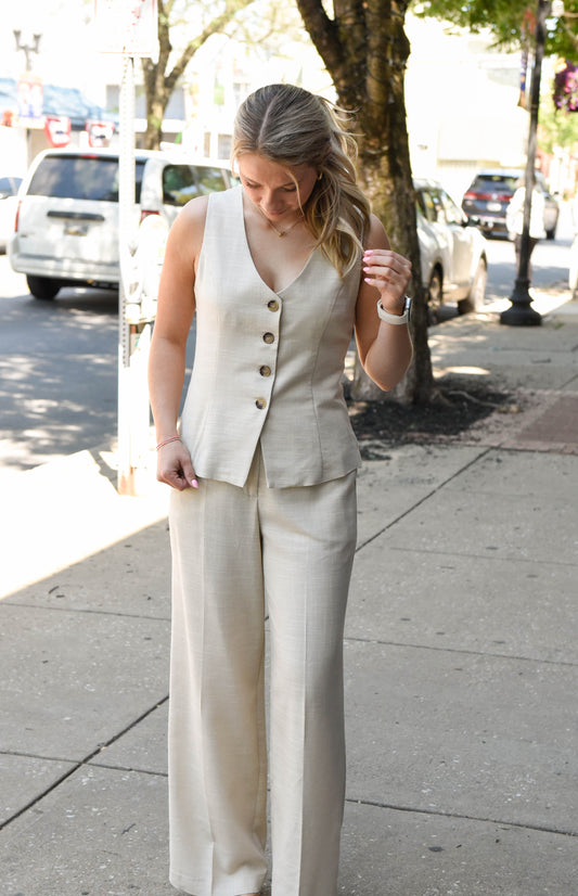 Woman in a beige vest and pants standing on a city sidewalk.