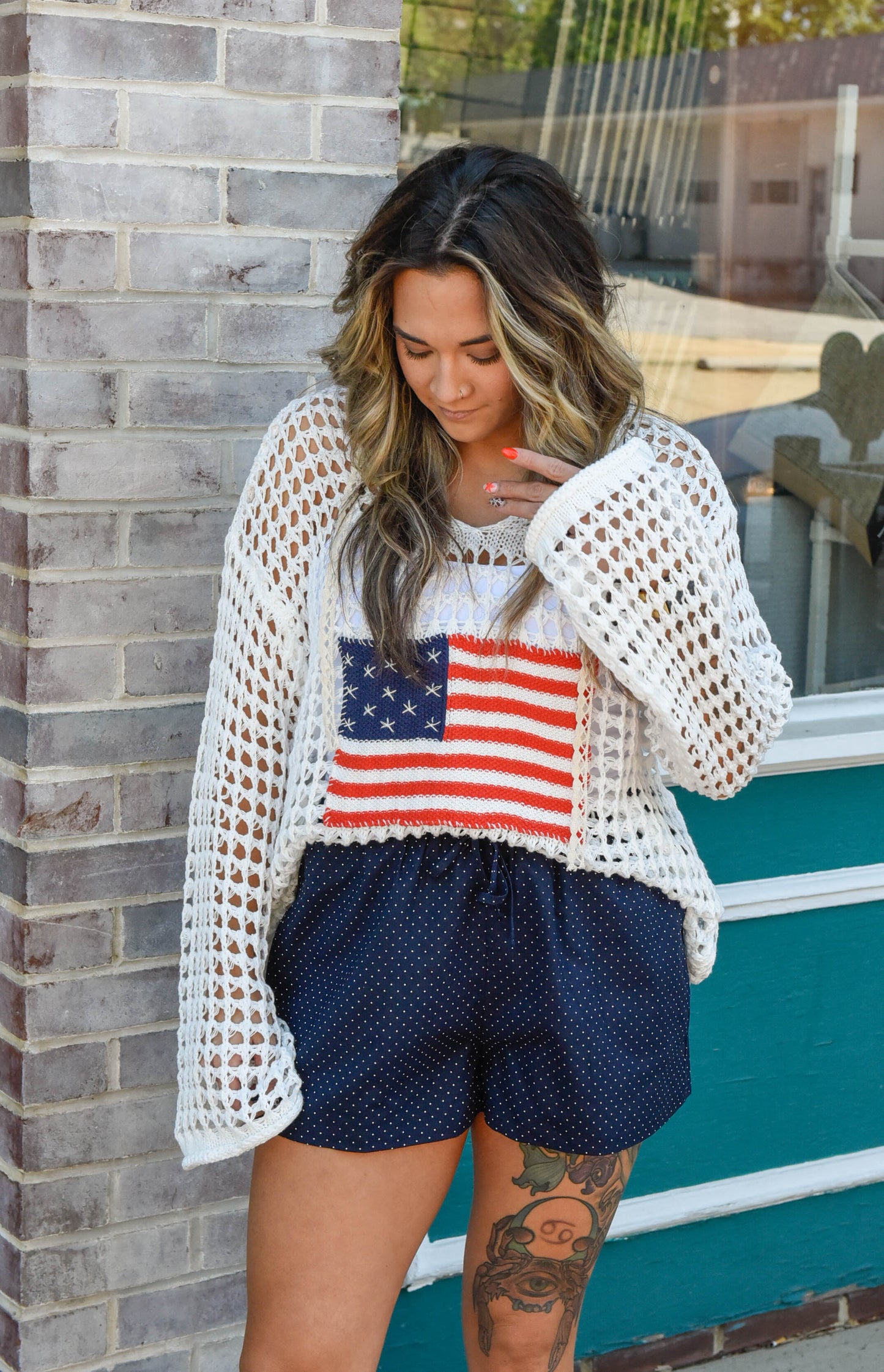 Woman wearing a white crochet cover-up with an American flag design, standing against a brick wall.