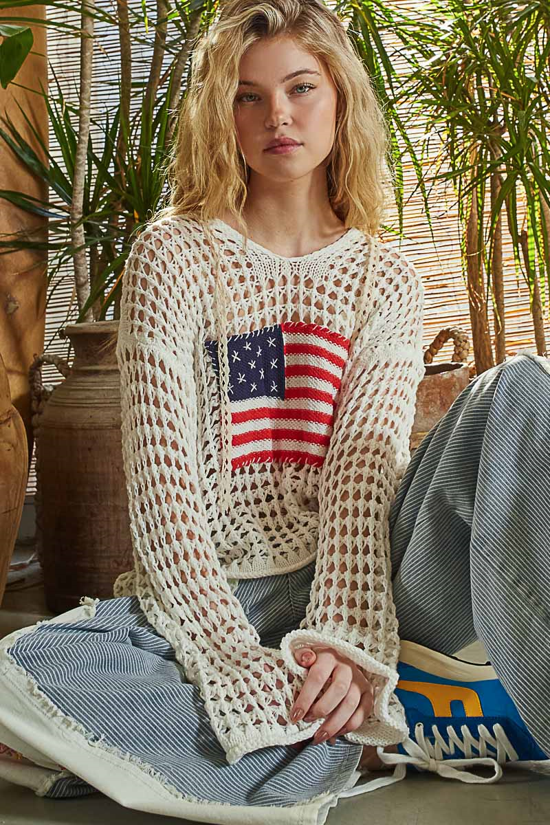 Woman wearing a white crochet sweater with an American flag design, sitting on the floor with plants in the background.