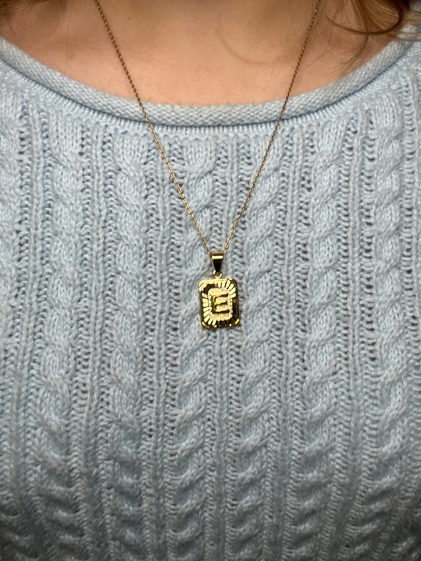 A gold initial necklace with a pendant on a person wearing a light blue sweater.