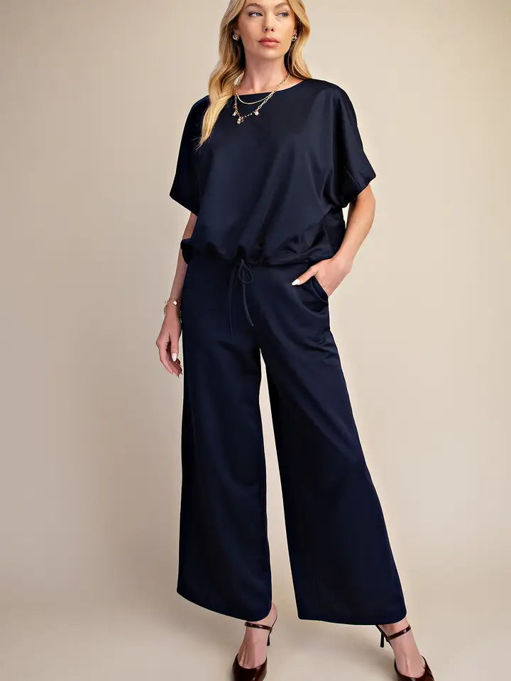 A woman modeling a navy blue satin jumpsuit with short flutter sleeves, a drawstring waist, and wide legs, paired with brown strappy heels against a solid beige studio background.