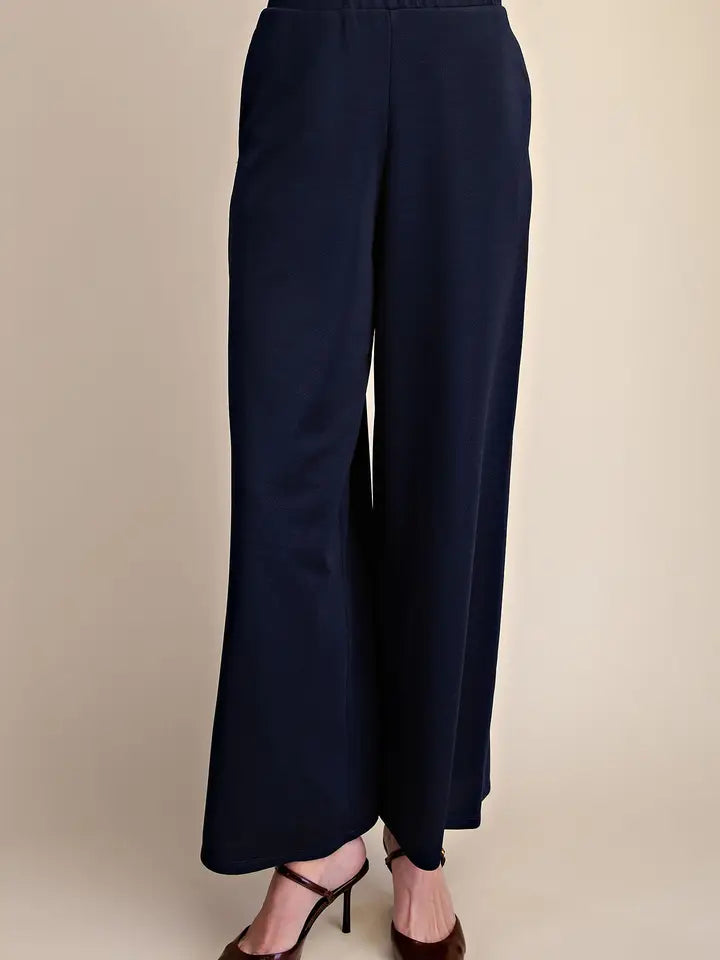 A person wearing dark navy wide-leg cropped pants with brown strappy high heels, shown from the waist down against a plain beige studio background.
