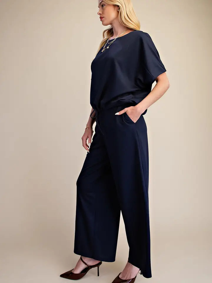 A woman modeling a navy blue short-sleeve jumpsuit with wide legs, standing against a plain beige studio background.