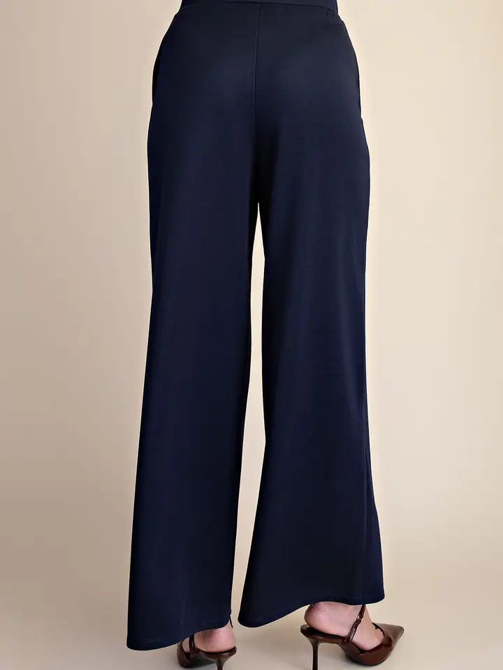 A person shown from the back wearing dark navy wide-leg pants and brown heeled sandals against a plain beige studio background.