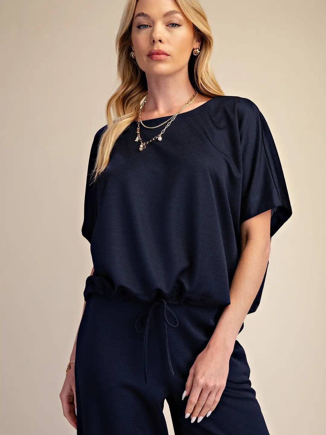 A woman modeling a navy blue short-sleeve top with a drawstring waist, paired with matching navy pants, against a solid beige background.