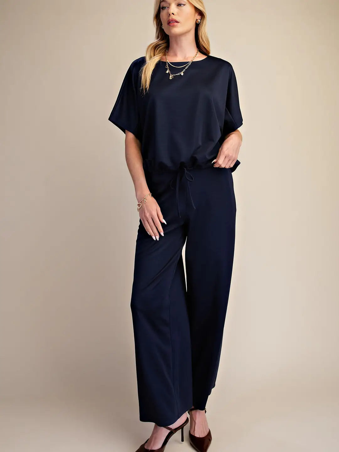 A woman modeling a navy blue two-piece set consisting of a short-sleeve top and matching wide-leg pants, styled with brown heels and layered gold necklaces against a solid beige studio background.