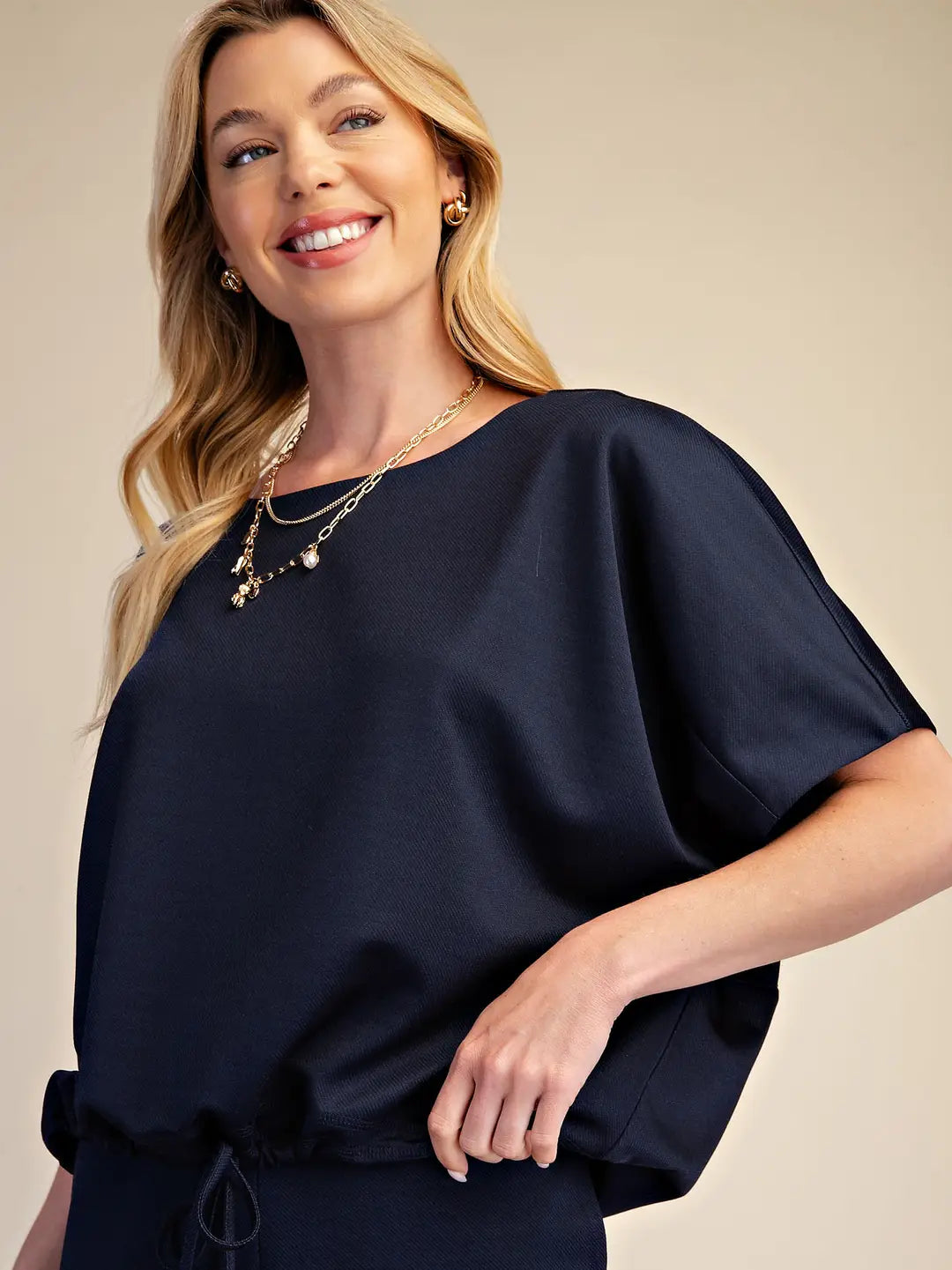 A smiling blonde woman wearing a loose, short-sleeve navy top with a drawstring waist and layered gold necklaces, posed against a plain beige studio background.