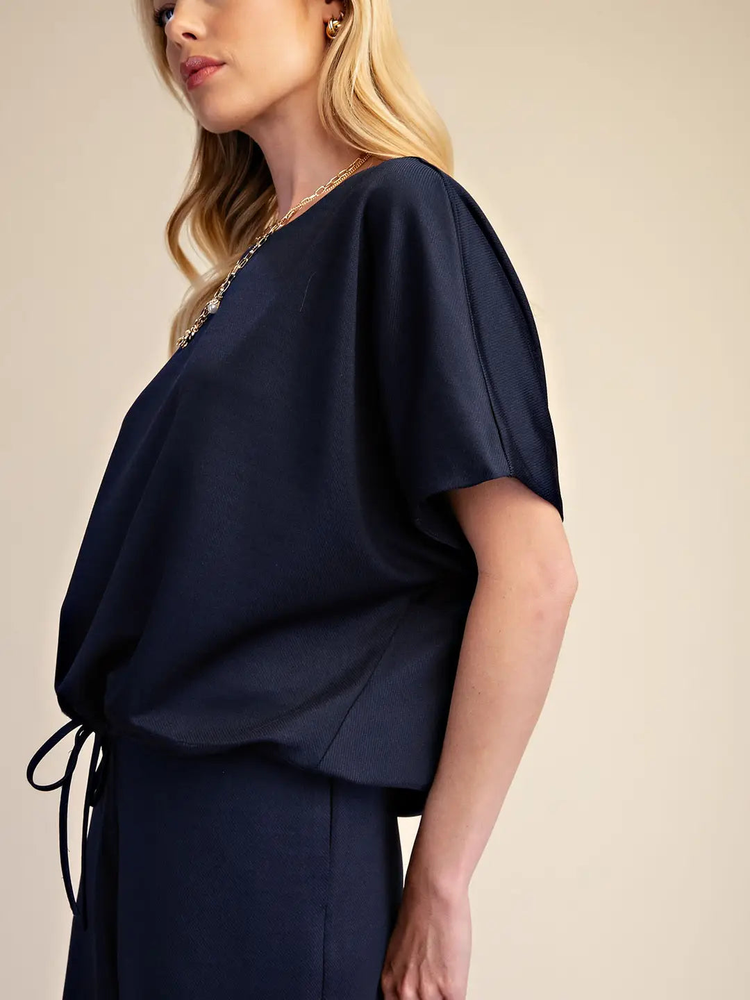 A woman wearing a navy blue short-sleeve top with a draped, oversized fit and a side tie detail, paired with matching navy bottoms, against a plain beige background.