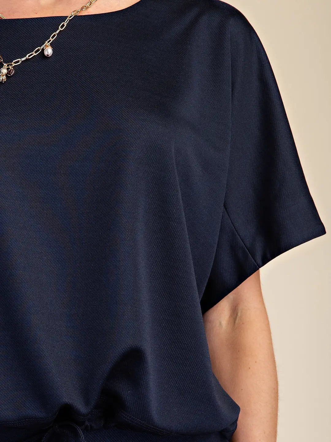 A close-up of a person wearing a dark navy blue short-sleeve top with a relaxed, draped fit. The image shows the chest, shoulder, arm, and a portion of the necklace against a solid beige background.