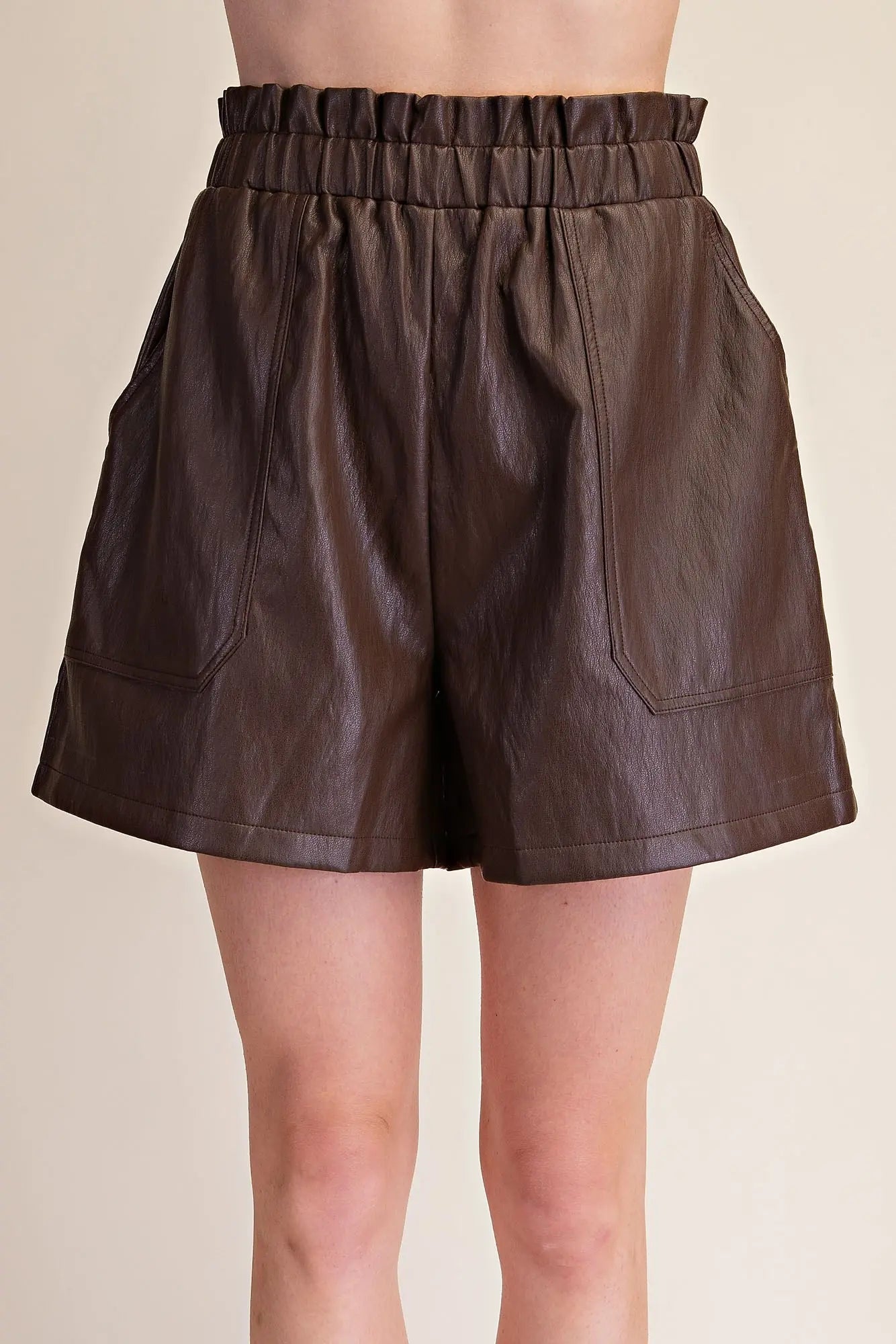 A person wearing dark brown faux-leather shorts with an elastic gathered waistband and large front patch pockets, shown against a plain light beige studio background.