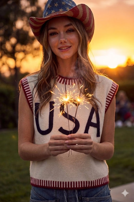 Woman wearing a 'USA' sweater and American flag hat holding sparklers at sunset.
