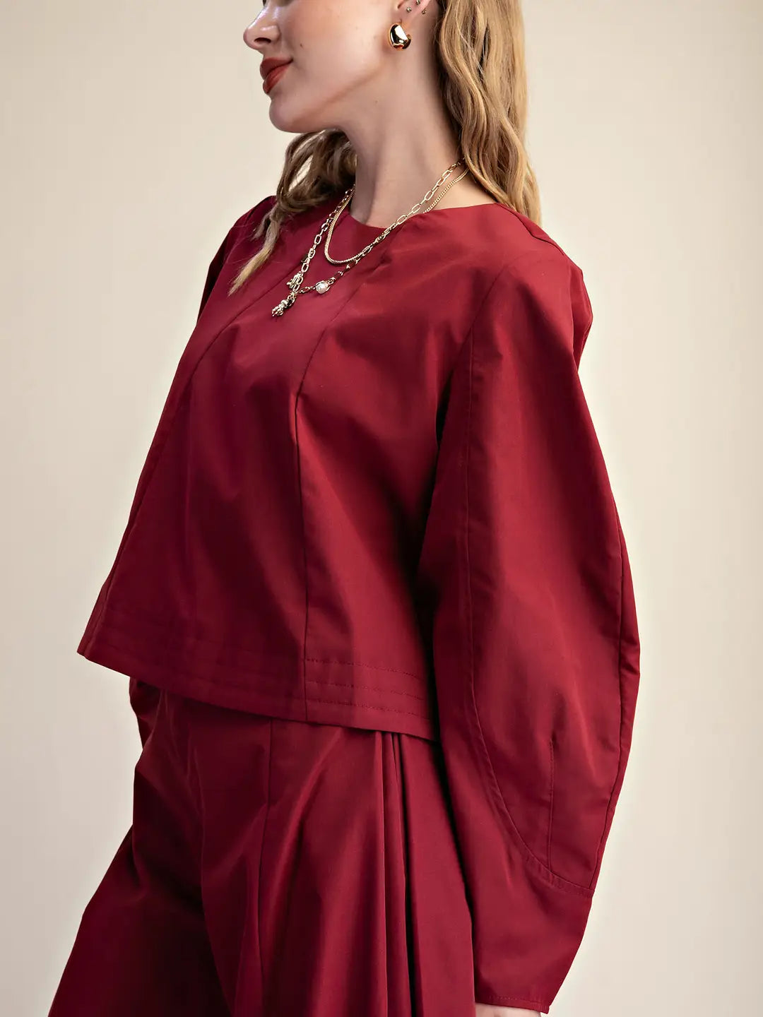 A woman wearing a matching deep red two-piece outfit with a cropped long-sleeve top and pleated pants, styled with layered gold necklaces and earrings against a plain beige background.