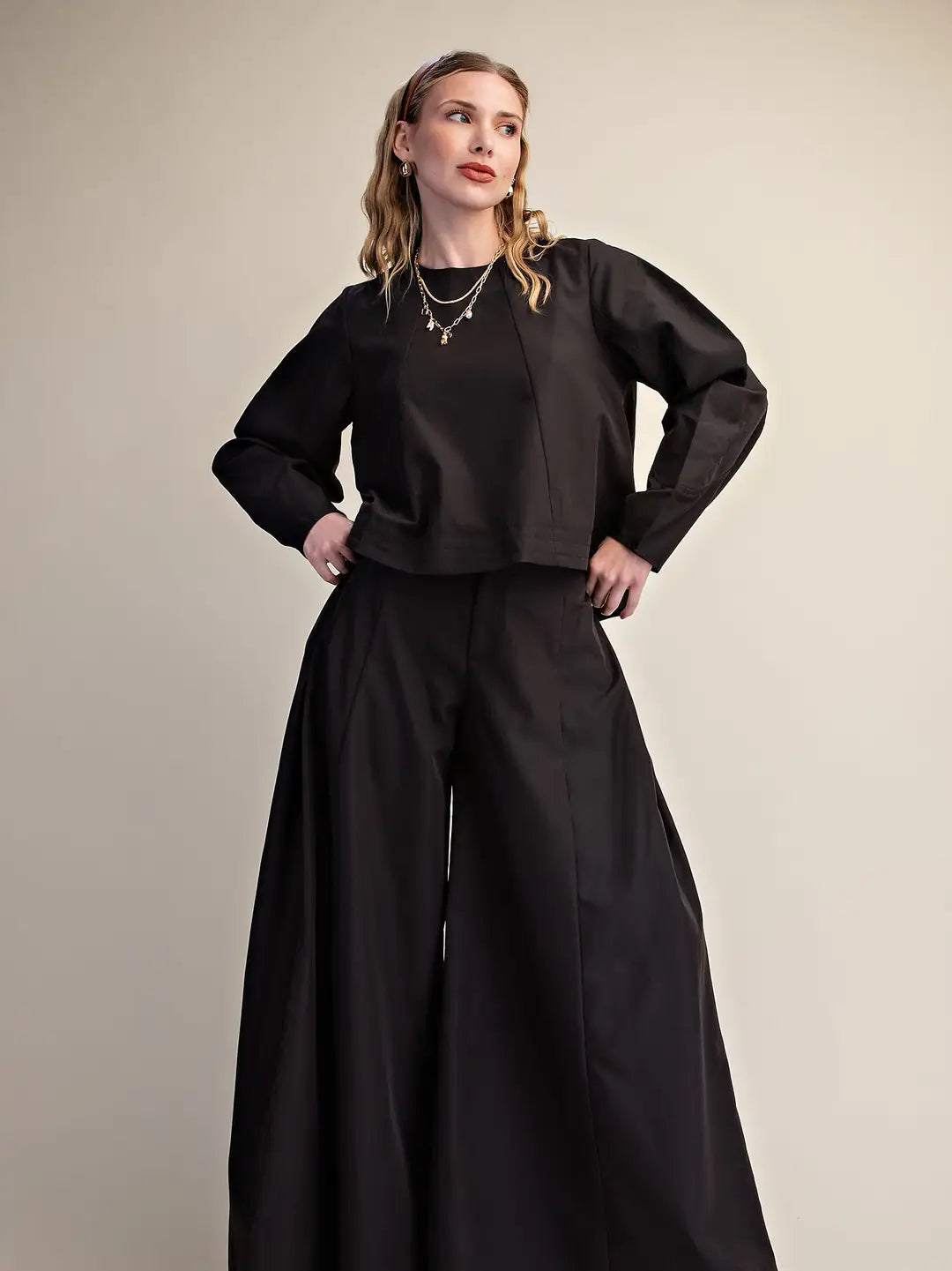 A woman modeling a black long-sleeve cropped top with structured shoulders and wide-leg black trousers, standing against a plain beige studio background.