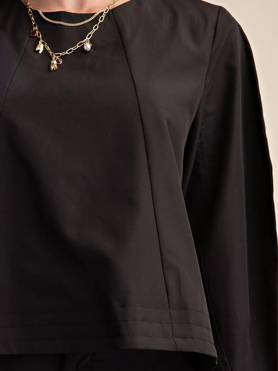 Close-up of a person wearing a black long-sleeve top with a relaxed, draped fit and a gold chain necklace with small charms, against a light neutral background.