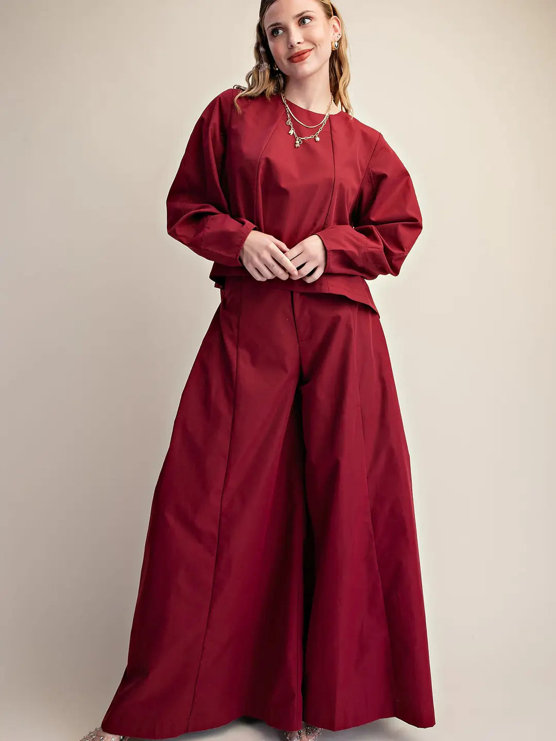 A woman modeling a matching two-piece outfit consisting of a long-sleeve cropped top and wide-leg trousers in a deep burgundy red, set against a plain beige studio background.