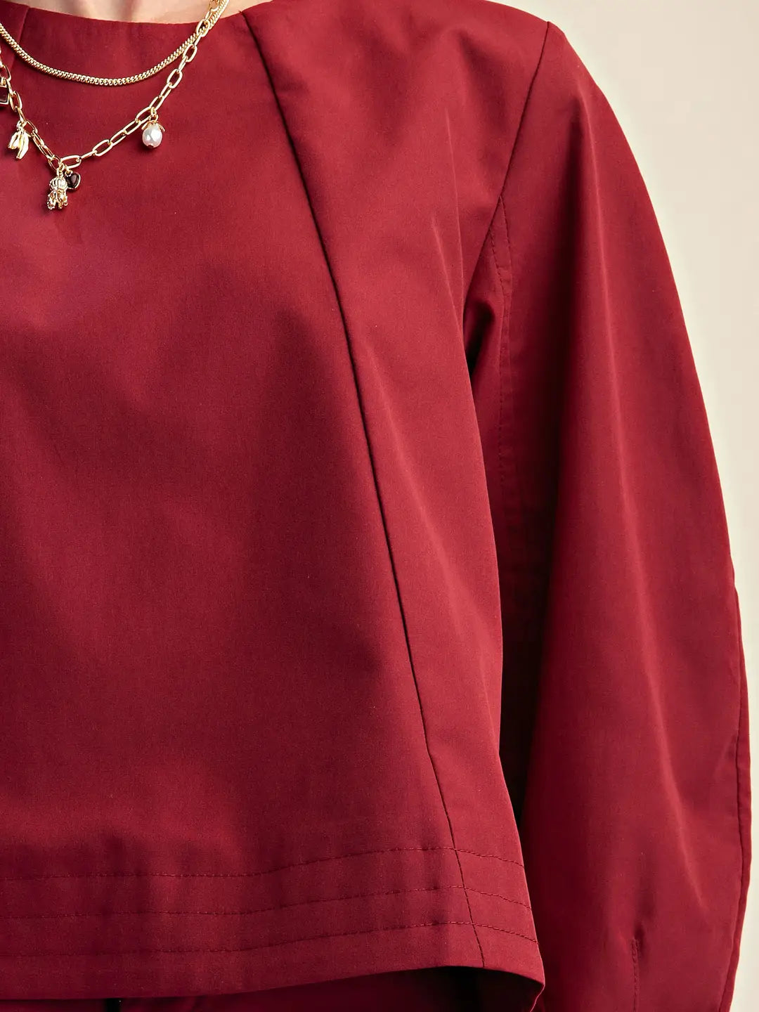 A close-up of a person wearing a long-sleeve top in deep burgundy red, featuring a high neckline and a delicate gold chain necklace with small charms.