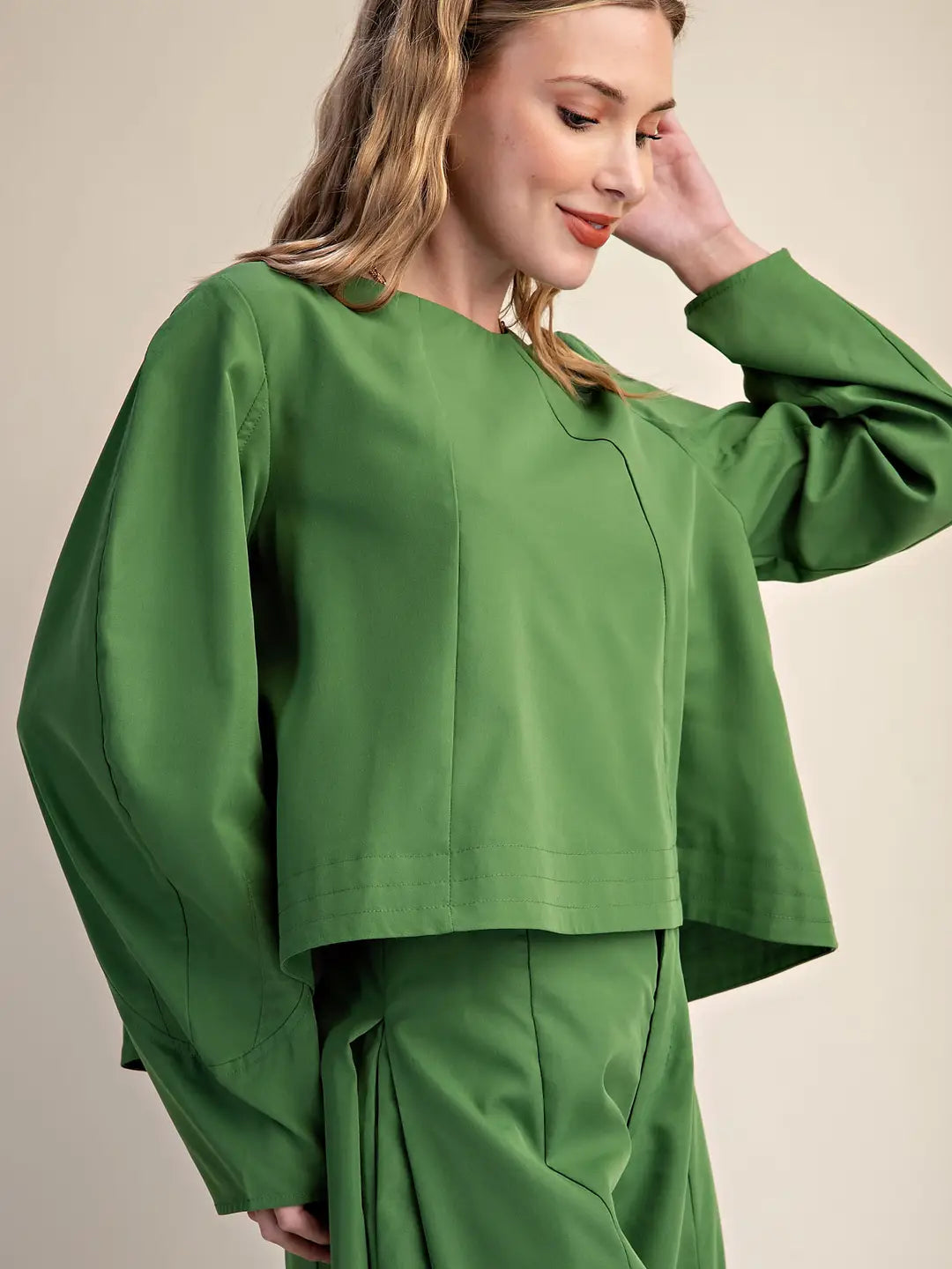 A woman wearing a vibrant green long-sleeve cropped top and matching green trousers, posing against a neutral beige background.