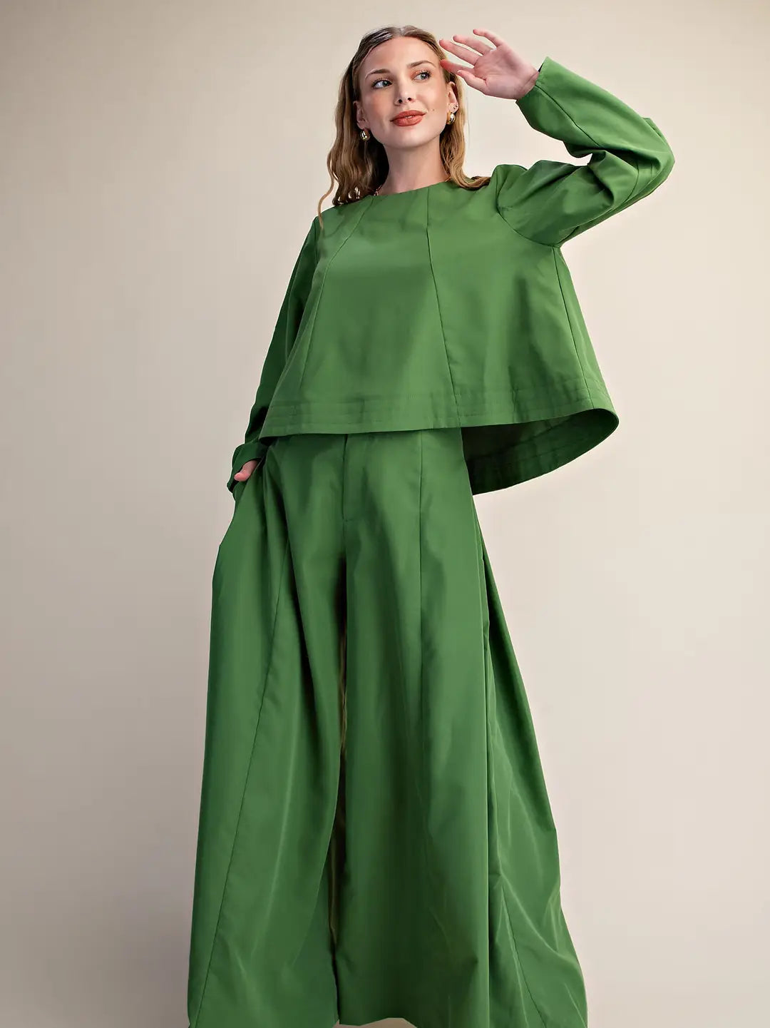 A woman modeling a matching green two-piece outfit consisting of a long-sleeve cropped top and wide-leg trousers, set against a plain beige studio background.