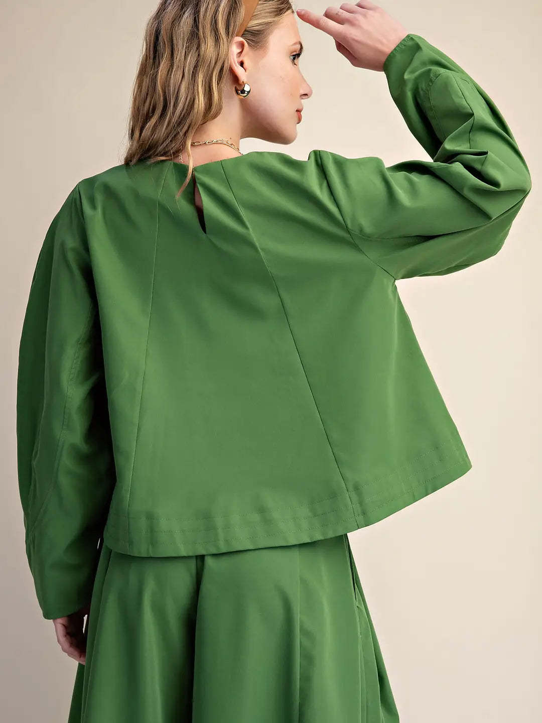 A woman shown from the back wearing a bright green cropped long-sleeve top and matching green skirt against a plain beige studio background.