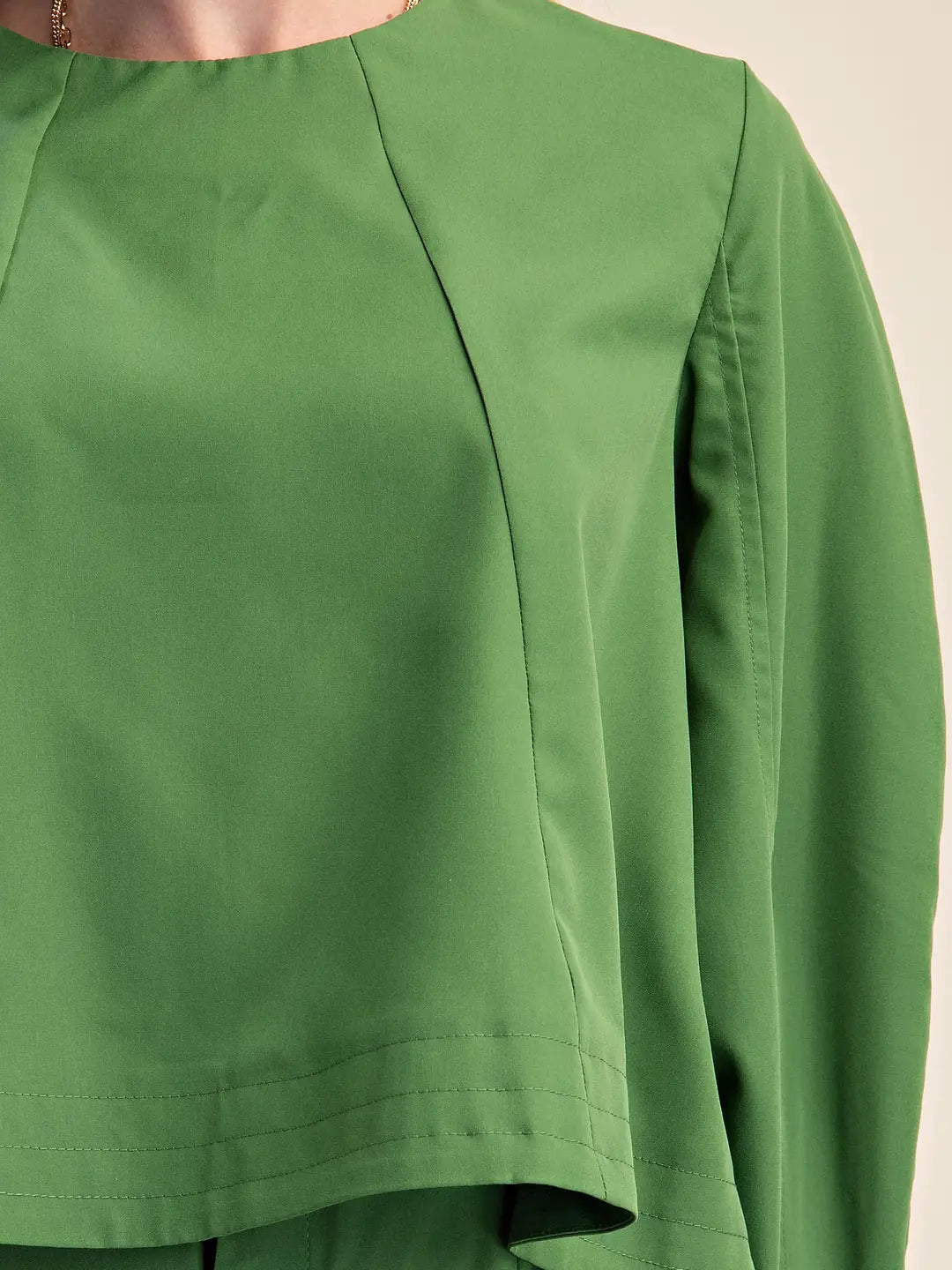 Close-up of a green long-sleeve top with a high-low hemline and structured shoulder seams, shown on a model against a light beige background.