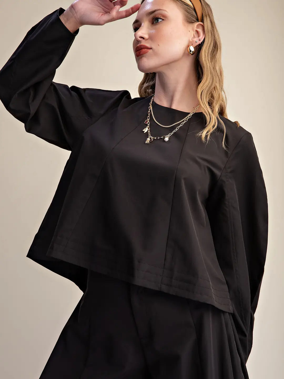 A woman wearing a black long-sleeve cropped top with a relaxed, draped silhouette and matching black bottoms, styled with layered gold necklaces and a tan headband against a plain beige studio background.