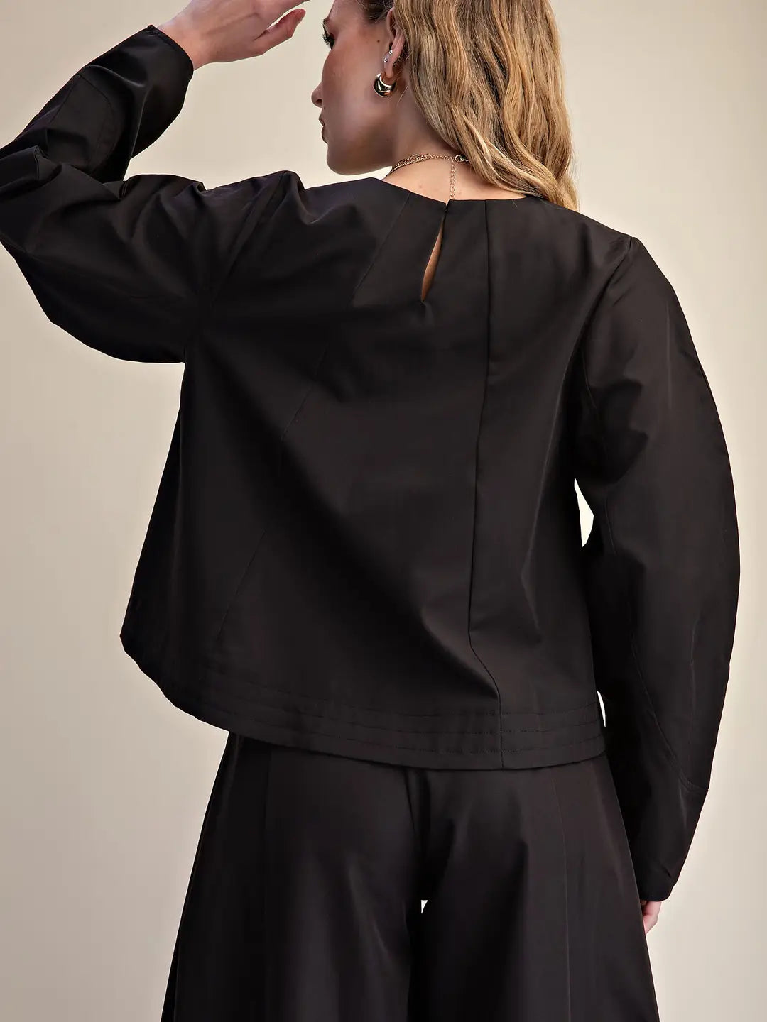 A woman shown from the back wearing a black long-sleeve cropped top and matching black bottoms against a plain beige studio background.