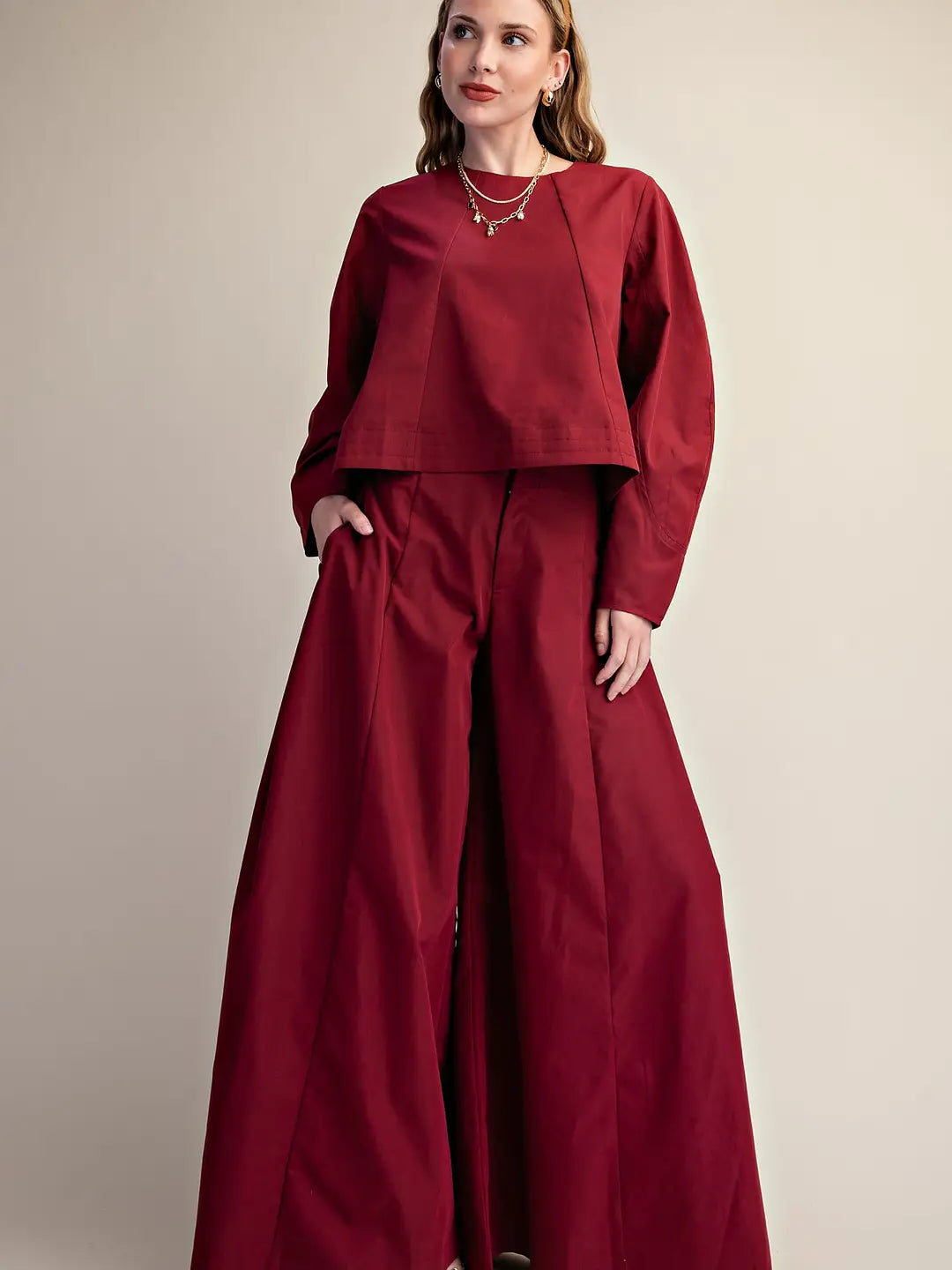 A woman modeling a matching deep red two-piece outfit consisting of a cropped long-sleeve top with draped shoulders and high-waisted wide-leg trousers, set against a plain beige studio background.