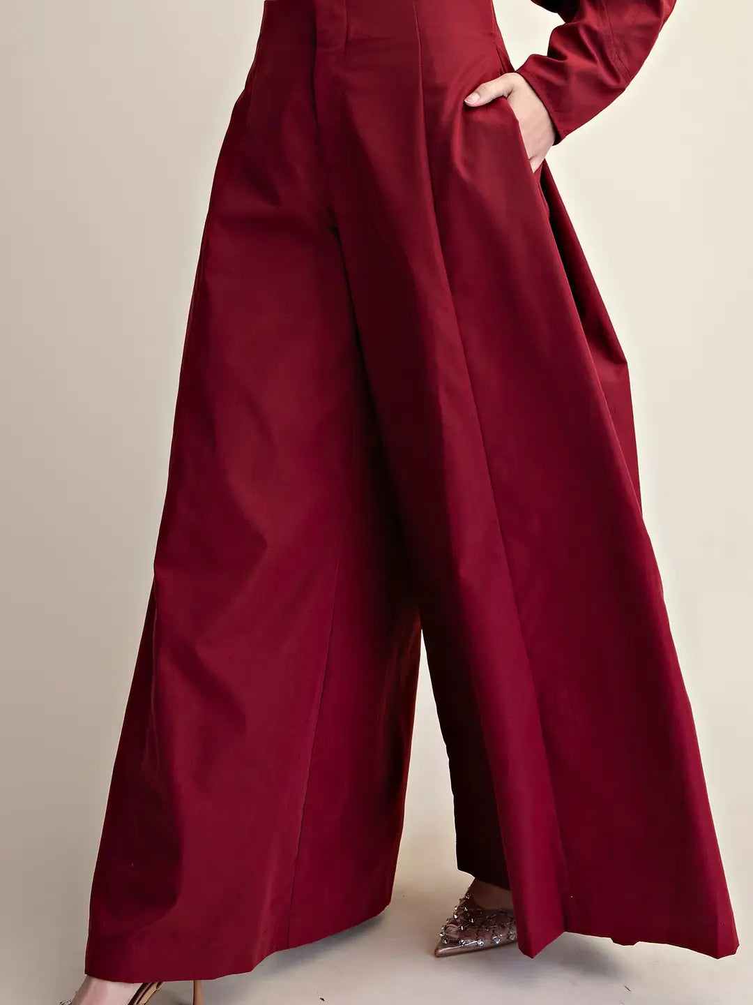 A close-up of a person wearing high-waisted, wide-leg trousers in a deep burgundy red fabric. The pants feature a high waist, front pleats, and side pockets, with one hand tucked into the pocket.
