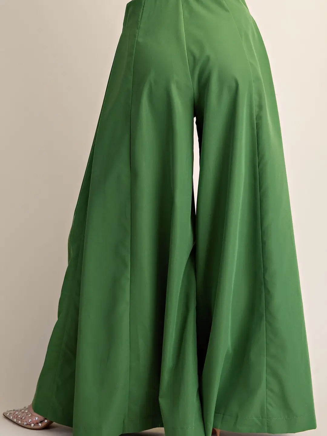 Green wide-leg pants shown from the back, featuring a high waist and a flowing, draped silhouette, with a pair of silver studded flat shoes partially visible at the bottom left.