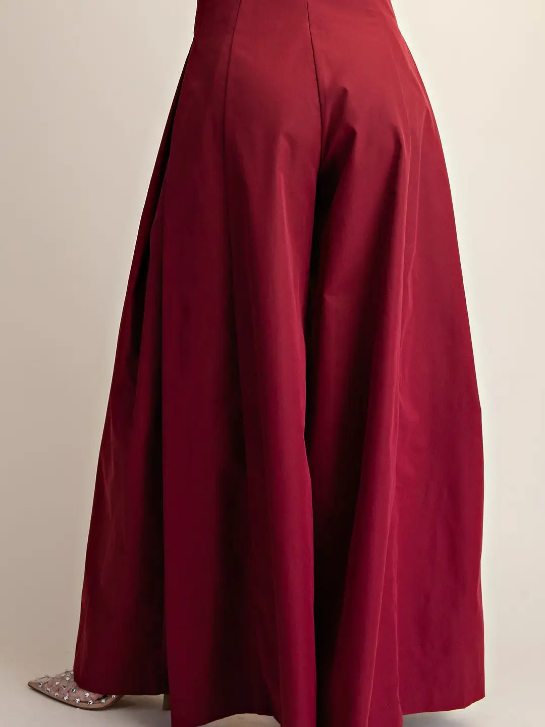 A close-up of a person wearing a voluminous, floor-length pleated skirt in deep burgundy red, paired with sparkly silver pointed flats, against a plain light beige background.