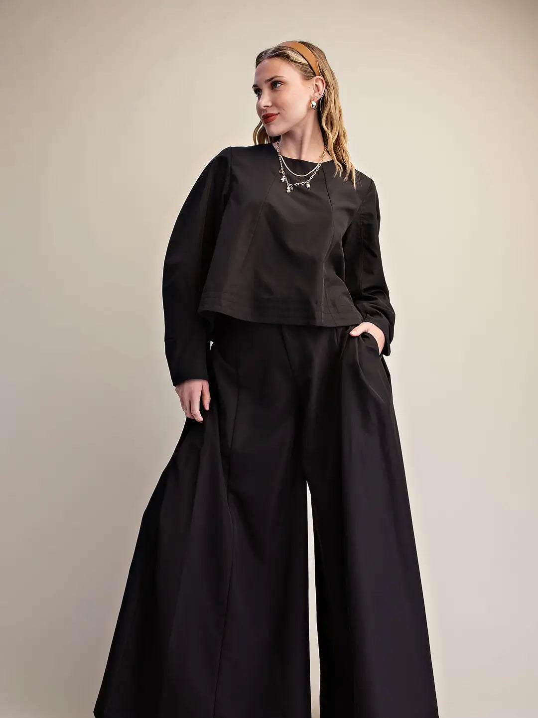A woman modeling a black long-sleeve cropped top and matching wide-leg trousers against a plain beige studio background.