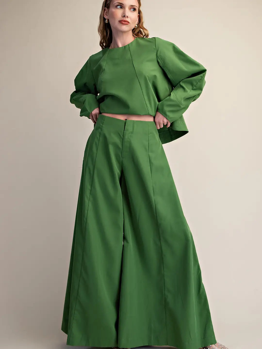 A woman modeling a matching green two-piece outfit consisting of a cropped long-sleeve top with dramatic puffed sleeves and high-waisted wide-leg pants, photographed against a plain beige studio background.