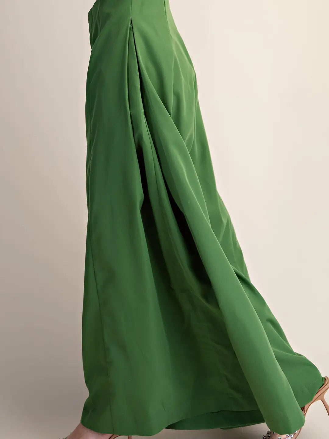 A close-up of a person wearing a flowing green skirt or drape of fabric, shown against a plain light beige studio background. The lower body and part of the feet are visible, and the fabric is styled as if being worn.
