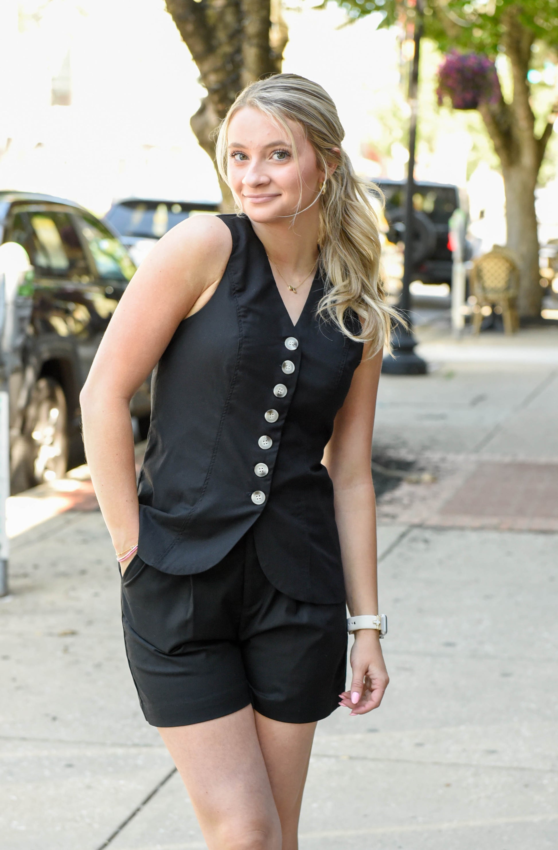 Woman wearing a black sleeveless top with buttons and shorts on a city street.