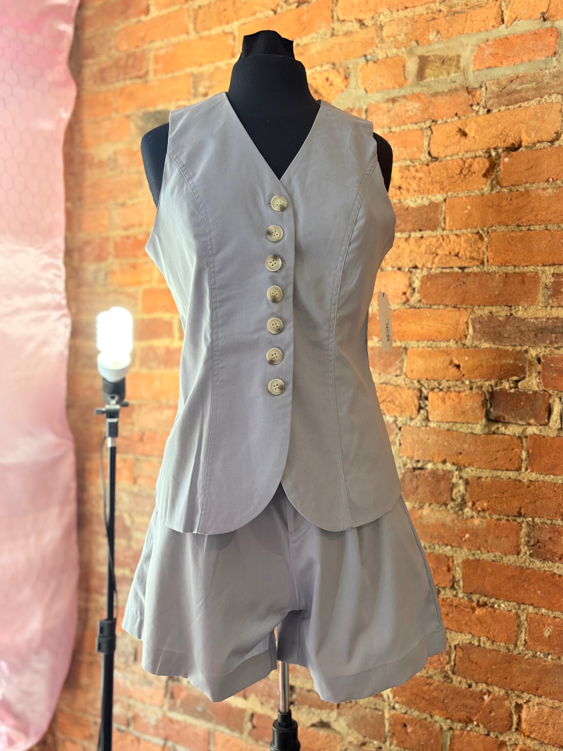 Light gray sleeveless top with buttons and shorts on a mannequin against a brick wall.