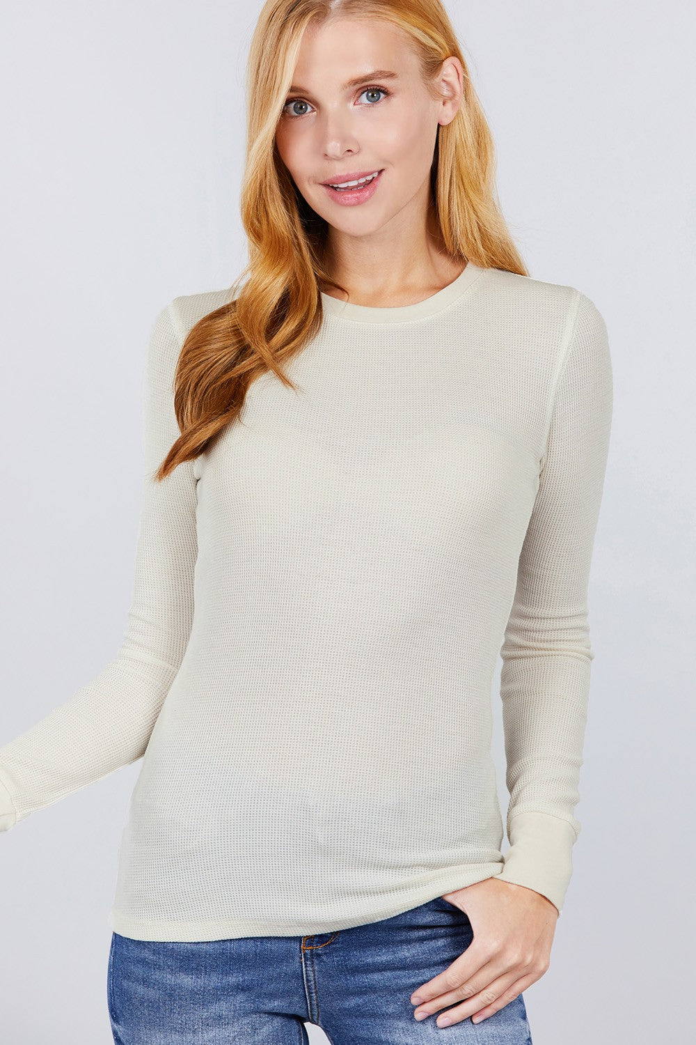 A woman with blonde hair wearing a light beige long-sleeve crewneck knit sweater and blue jeans, photographed against a light gray studio background.