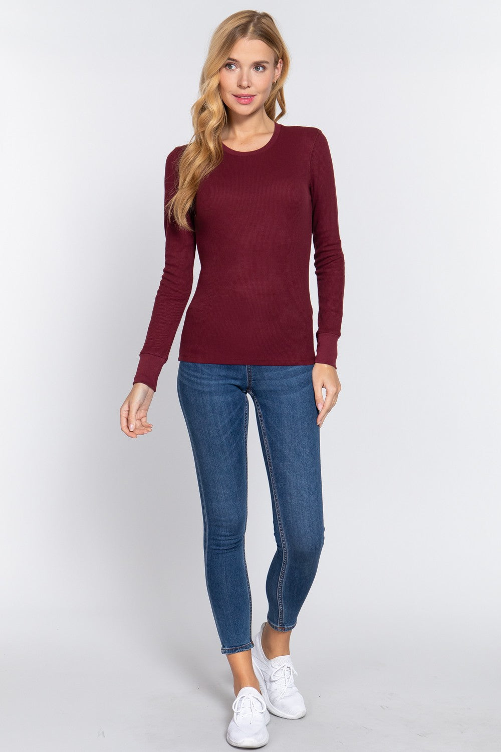 A woman modeling a long-sleeve crewneck knit top in burgundy, paired with blue skinny jeans and white sneakers, against a light gray studio background.