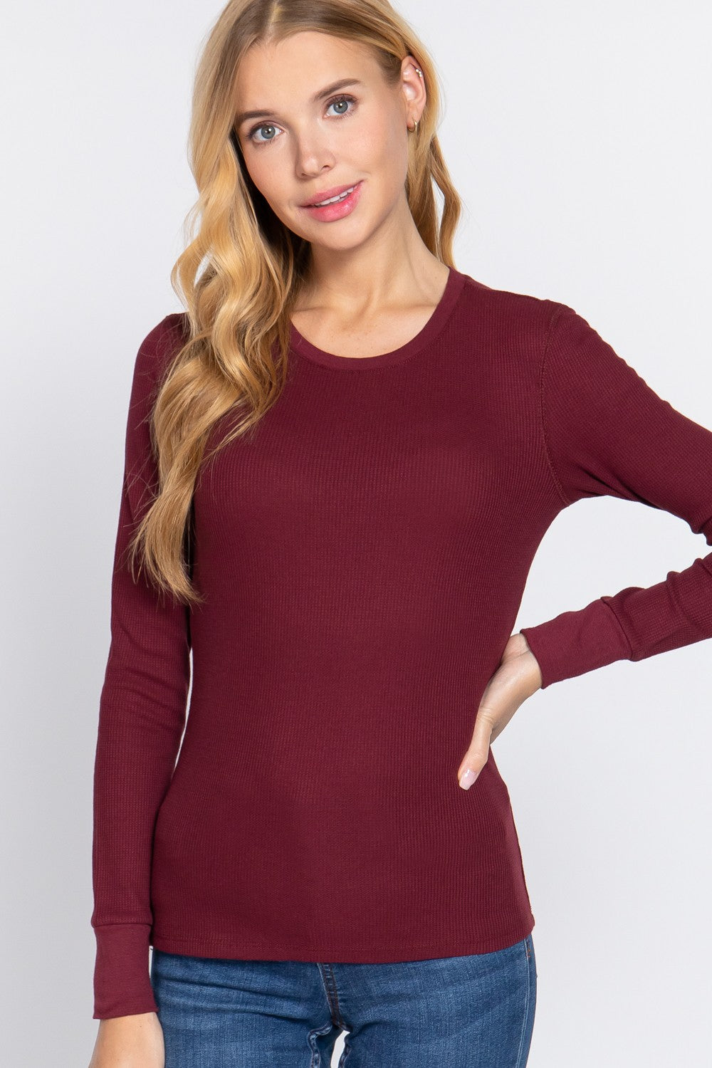 A woman with blonde hair wearing a long-sleeve crewneck knit top in burgundy, paired with blue jeans, against a light gray studio background.