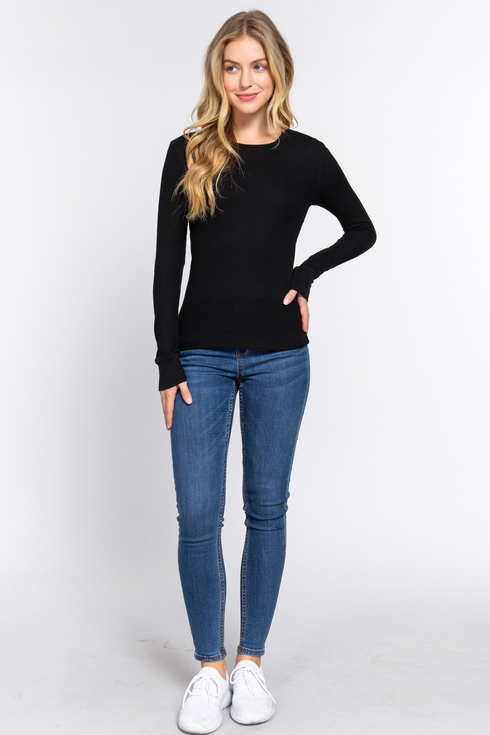 A woman modeling a black long-sleeve crewneck sweater, blue skinny jeans, and white sneakers against a plain white studio background.