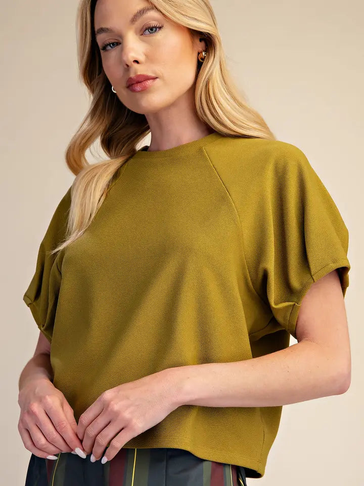 A blonde woman wearing a short-sleeve olive green top with a relaxed, slightly cropped fit and gathered sleeves, paired with dark patterned trousers, set against a plain beige studio background.