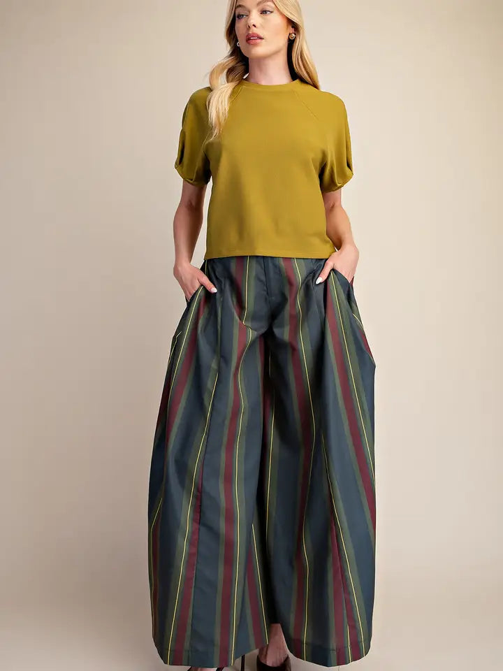 A woman modeling a short-sleeve olive green top paired with wide-leg dark gray trousers featuring vertical red, green, and yellow stripes, set against a plain beige studio background.