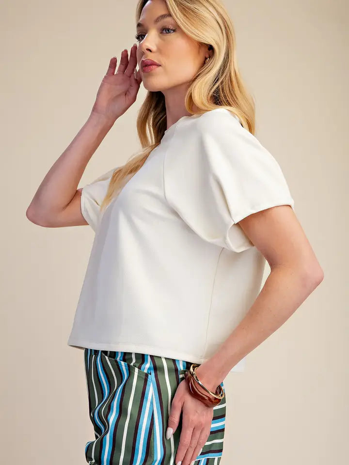 A blonde woman wearing a white short-sleeve crop top with ruffled shoulders and striped green, blue, and white trousers, posing against a solid beige background.