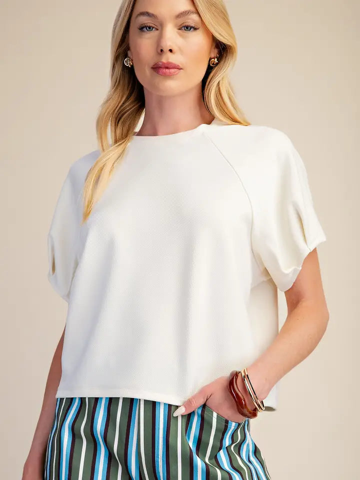 A blonde woman wearing a white short-sleeve top with raglan sleeves and striped green, blue, and white trousers, photographed against a warm beige studio background.